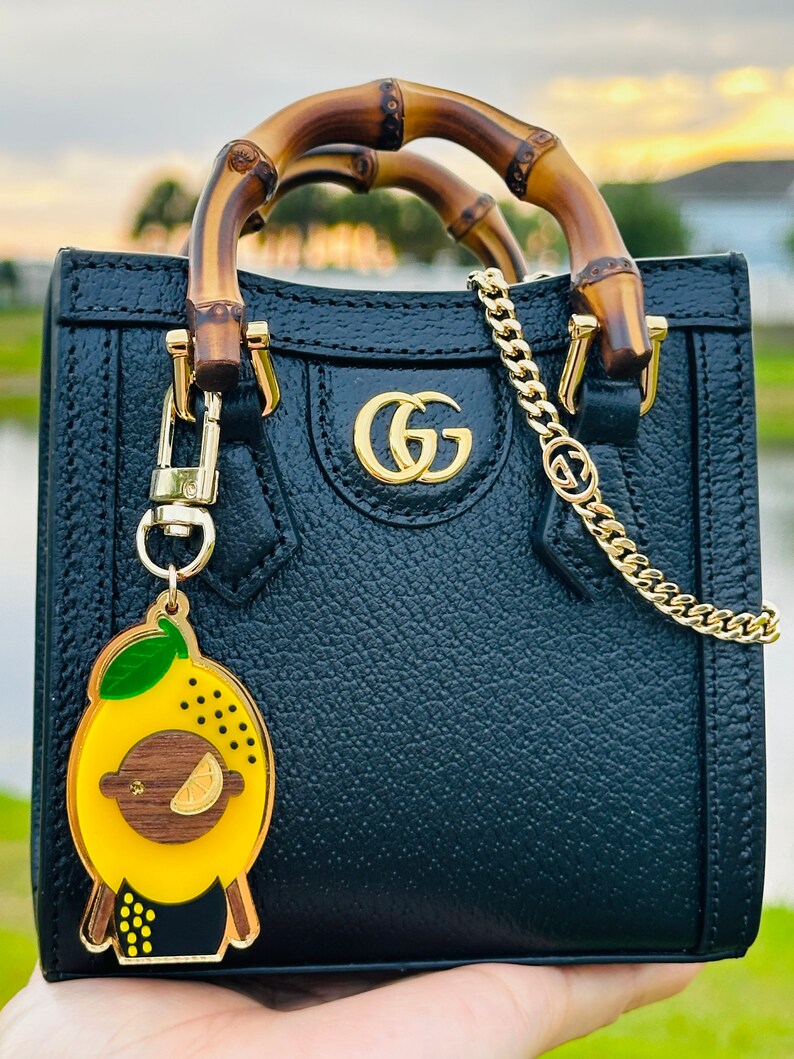 May include: Black leather handbag with bamboo handles and a gold chain strap. The bag has a gold GG logo on the front and a lemon-shaped keychain.