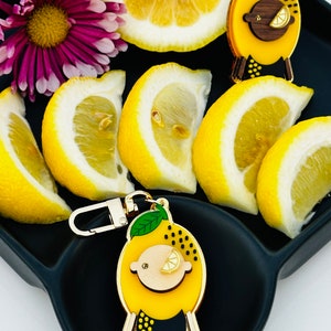 May include: A gold-tone keychain charm shaped like a cartoon lemon with a black and yellow patterned body, green leaves, and a gold-tone face with a small lemon slice for an eye. The charm is on a black tray with lemon slices.