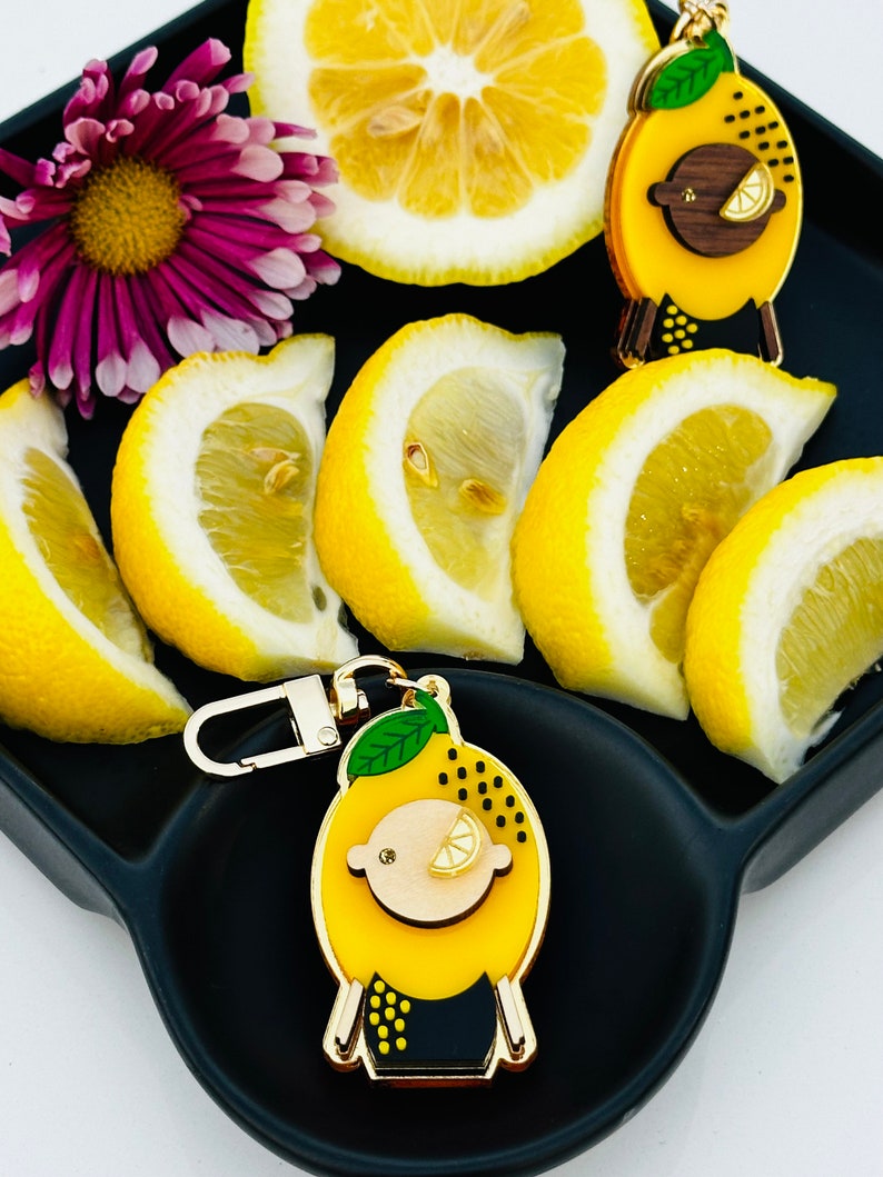 May include: A gold-tone keychain charm shaped like a cartoon lemon with a black and yellow patterned body, green leaves, and a gold-tone face with a small lemon slice for an eye. The charm is on a black tray with lemon slices.