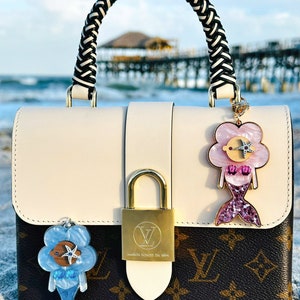 May include: A white and brown designer handbag with a braided black and white handle. The bag has a gold lock and two charms attached to it. The charms are blue and pink mermaids with glitter accents. The bag is sitting on a sandy beach.