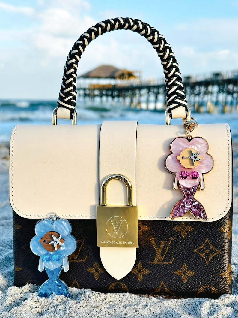 May include: A white and brown designer handbag with a braided black and white handle. The bag has a gold lock and two charms attached to it. The charms are blue and pink mermaids with glitter accents. The bag is sitting on a sandy beach.