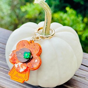 May include: A small, orange and brown resin keychain shaped like a person with a green leaf on their head. The keychain is attached to a gold key ring and is hanging from a white pumpkin.
