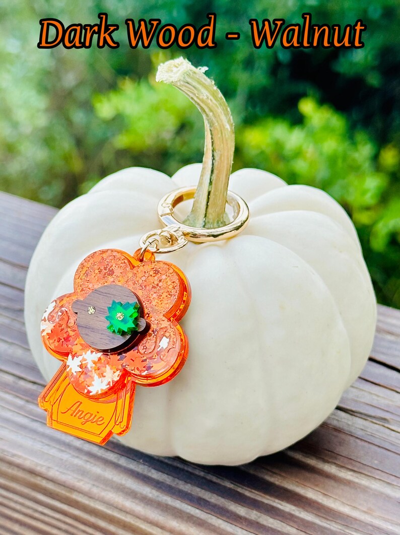 May include: A small, orange and brown resin keychain shaped like a person with a green leaf on their head. The keychain is attached to a gold key ring and is hanging from a white pumpkin.