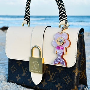 May include: A white and brown monogrammed handbag with a black and white braided handle. The bag has a gold lock and a pink and white mermaid charm attached to it. The bag is sitting on a sandy beach.