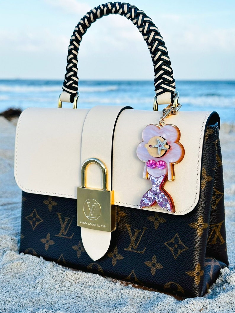 May include: A white and brown monogrammed handbag with a black and white braided handle. The bag has a gold lock and a pink and white mermaid charm attached to it. The bag is sitting on a sandy beach.