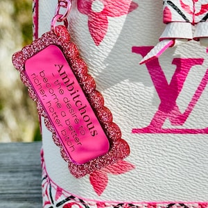May include: A pink glittery charm with the word "Ambitchous" and the phrase "the desire to become a better richer, hotter bitch" is attached to a white and pink patterned handbag. The handbag has a pink floral design.