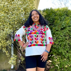 Op de afbeelding: Een wit geborduurd blouse met kleurrijke bloemen- en geometrische patronen. De blouse heeft een ronde hals en driekwart mouwen. Het model draagt een zwarte short.