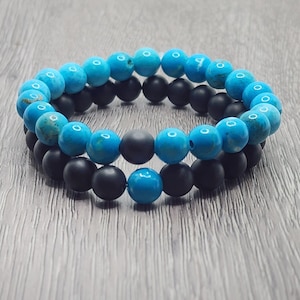 Könnte beinhalten: Zwei Armbänder mit Perlen. Ein Armband hat türkisfarbene Perlen, das andere schwarze, matte Perlen. Die Armbänder sind übereinander gestapelt. Der Hintergrund ist eine Holzoberfläche.