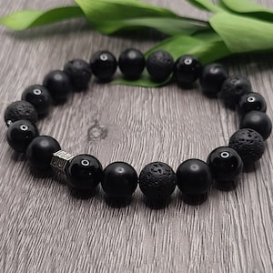 Könnte beinhalten: Ein schwarzes Perlenarmband mit abwechselnden schwarzen Lavasteinen und Onyxperlen. Das Armband hat einen silbernen Anhänger mit einem geometrischen Muster.