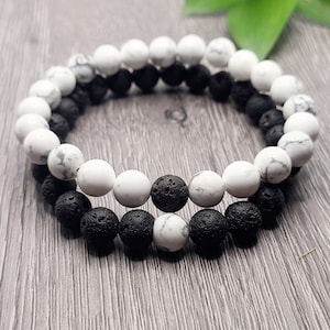 Könnte beinhalten: Zwei Armbänder, eines mit weißen Perlen und eines mit schwarzen Lava-Perlen. Die Armbänder sind übereinander gelegt.