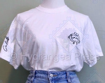 Puede incluir: Camiseta blanca con un personaje de manzana de dibujos animados en blanco y negro en la parte delantera y el texto "Caleb" en la manga izquierda. La camiseta está puesta en una persona que lleva pantalones vaqueros azules.