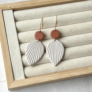 May include: A pair of white clay earrings with a textured leaf design and a brown clay accent. The earrings are hanging from gold hoops.