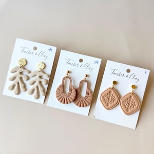 May include: Three pairs of handmade clay earrings displayed on white cards. The earrings feature various designs: leaf-shaped, arched with sunburst details, and diamond-shaped with floral patterns. Each pair has a gold-colored stud.