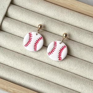 May include: A pair of dangle earrings featuring circular white charms painted with red baseball stitching patterns. Each charm is suspended from a small gold-toned metal ball stud post, creating a simple and distinct sports-themed accessory.
