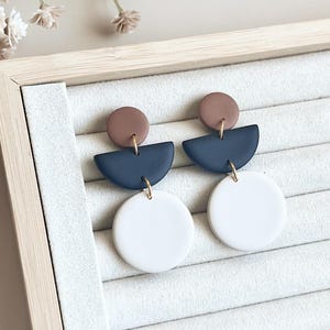 May include: A pair of modern geometric earrings. Each earring features a small brown circle, a navy blue half-circle, and a large white circle, all connected with gold-colored metal findings. The earrings are displayed on a light-colored, textured surface.