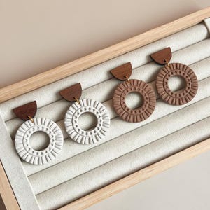 May include: Four pairs of handcrafted earrings displayed in a wooden jewelry box. Each earring features a half-circle wooden top and a circular pendant with a textured design. The pendants are in white and brown colors.