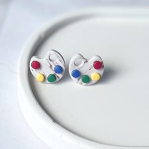 May include: A pair of artistic earrings shaped like paint palettes. Each earring is white with brown marbling and features four colorful paint dots: red, blue, green, and yellow. The earrings are set against a white background.
