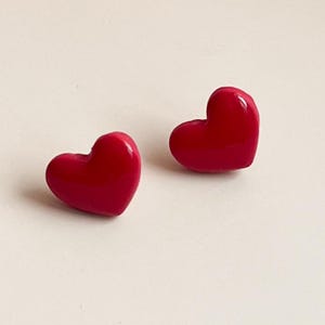 May include: Two glossy, heart-shaped stud earrings in a vibrant red hue. The earrings are positioned on a light-colored surface, showcasing their smooth, rounded forms. These earrings are a simple yet charming accessory.