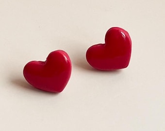 May include: Two glossy, heart-shaped stud earrings in a vibrant red hue. The earrings are positioned on a light-colored surface, showcasing their smooth, rounded forms. These earrings are a simple yet charming accessory.