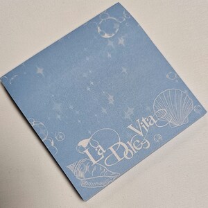 May include: A square, light blue sticky note with a design of bubbles, stars, and seashells. The words "La Dolce Vita" are written in a decorative font. The note is angled on a white surface.
