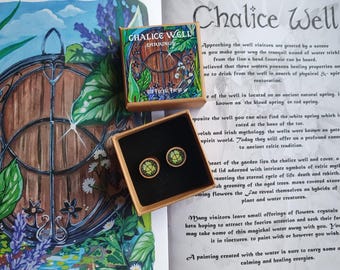May include: Wooden stud earrings with a green and gold design, presented in a small box with the text "Chalice Well Earrings". The earrings are displayed on a page with text about the Chalice Well.