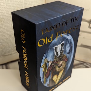 May include: A blue box with gold lettering that reads "Tarot of the Old Forest". The box features an illustration of a badger-headed person playing a lute.