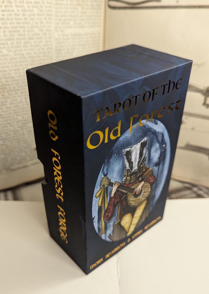 May include: A blue box with gold lettering that reads "Tarot of the Old Forest". The box features an illustration of a badger-headed person playing a lute.