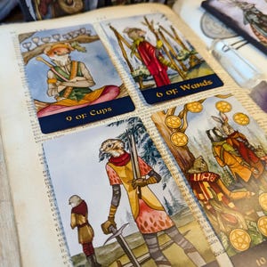 May include: Four tarot cards with illustrations of animals in a forest setting. The cards are 9 of Cups, 6 of Wands, 5 of Swords, and 10 of Pentacles. The cards are laid out on a book with a dark blue cover.