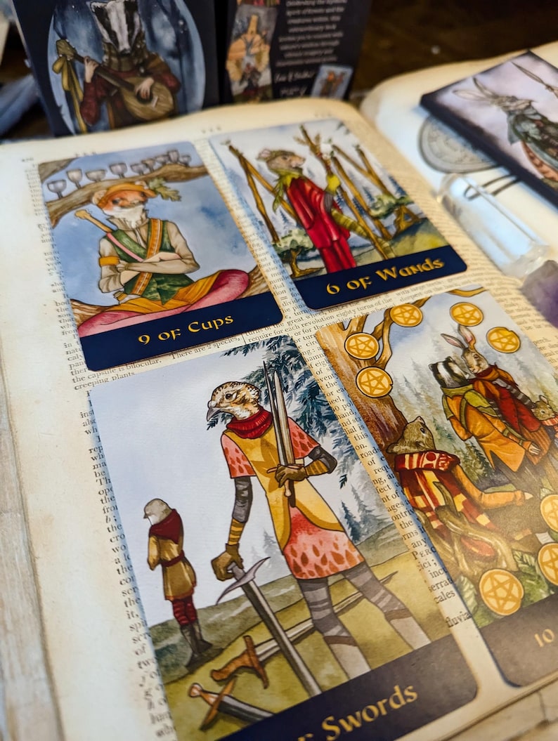 May include: Four tarot cards with illustrations of animals in a forest setting. The cards are 9 of Cups, 6 of Wands, 5 of Swords, and 10 of Pentacles. The cards are laid out on a book with a dark blue cover.