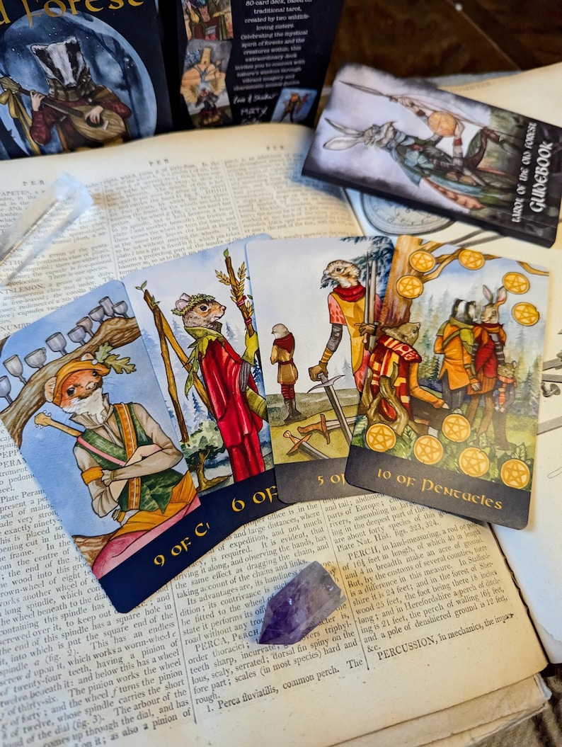 May include: A set of tarot cards featuring illustrations of woodland creatures. The cards show a badger, a squirrel, a rabbit, and a fox. The cards are numbered 9 of Cups, 6 of Swords, 5 of Swords, and 10 of Pentacles. The cards are on a book with a text about Perch.