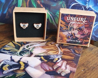 May include: Wooden bee earrings displayed in a small box with a black interior. The earrings are light brown with a bee design. A second box with the text "UNFURL EARRINGS" and an illustration is also present.