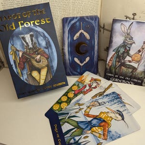 May include: Tarot of the Old Forest deck box with a badger playing a lute on the front. The box is blue with gold lettering. The cards are spread out on a table with a guidebook.