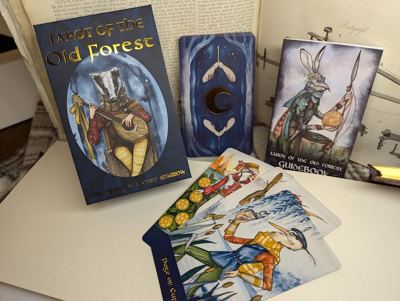 May include: Tarot of the Old Forest deck box with a badger playing a lute on the front. The box is blue with gold lettering. The cards are spread out on a table with a guidebook.