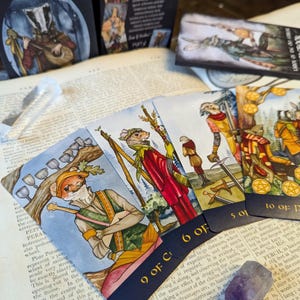 May include: A set of tarot cards featuring whimsical illustrations of animals in a forest setting. The cards are arranged in a fan shape, with the 9 of Cups, 6 of Cups, 5 of Cups, and 10 of Pentacles visible. The cards have a dark blue border with gold lettering.