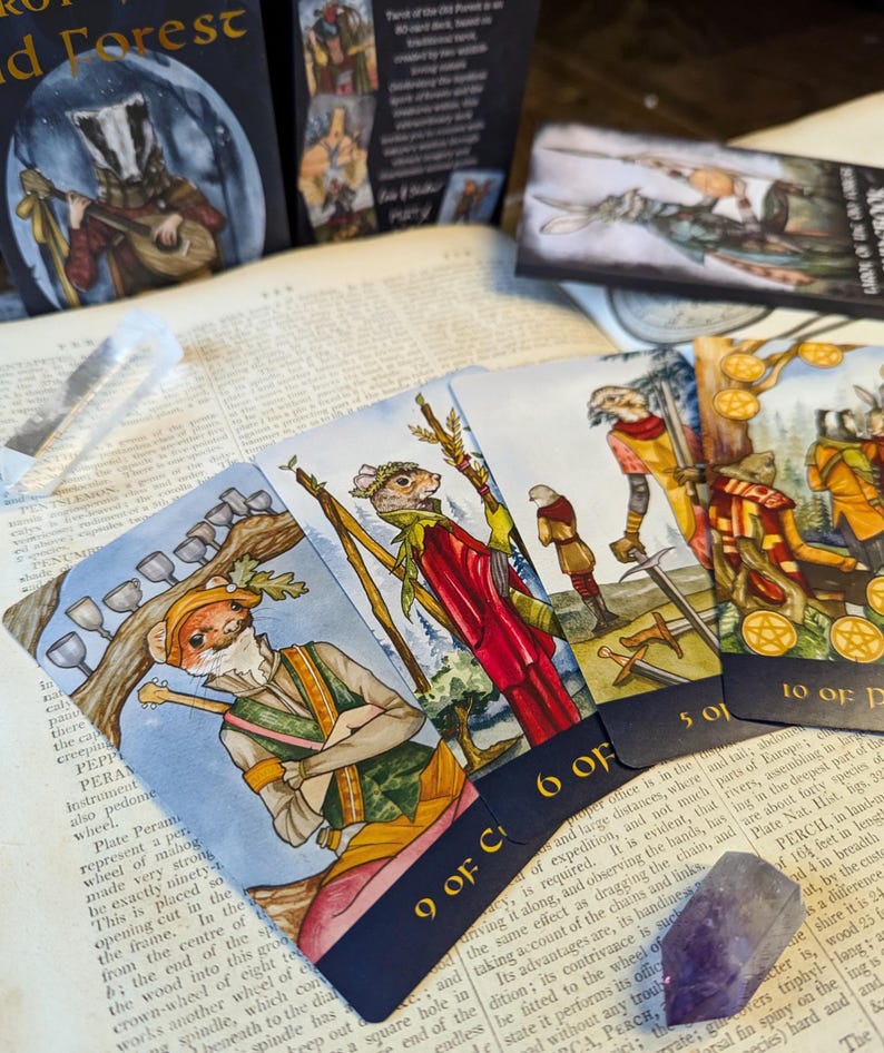 May include: A set of tarot cards featuring whimsical illustrations of animals in a forest setting. The cards are arranged in a fan shape, with the 9 of Cups, 6 of Cups, 5 of Cups, and 10 of Pentacles visible. The cards have a dark blue border with gold lettering.