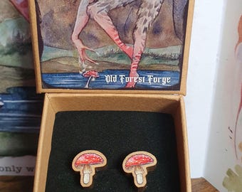 May include: A pair of wooden mushroom earrings with red caps and white stems, displayed in a brown box. The box lid features an illustration and the word "EARRINGS". The text "Old Forest Forge" is also visible.