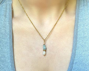 Könnte beinhalten: Eine zarte goldfarbene Halskette mit einem Anhänger aus hellblauen Steinen und einer weißen Perle. Die Halskette wird auf einem grauen Tanktop getragen. Die Kette hat ein gedrehtes Seildesign.