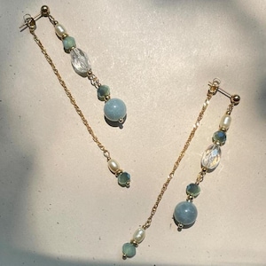 May include: A pair of gold-tone drop earrings featuring a post back, a long delicate chain, and a vertical arrangement of beads. The beads include white pearls, faceted clear crystals, and smooth light blue spheres, all connected by gold-tone metal.