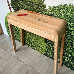 Peut inclure: Une console en bois clair avec un plateau rectangulaire et des bords arrondis. La table a un décor d'incrustation de petits carrés autour du bord du plateau et le long des pieds. La table est posée sur un sol en carrelage gris devant un mur vert.
