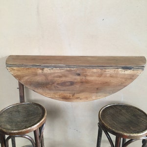 Peut inclure: Une table pliante en bois avec un plateau en demi-cercle, fixée au mur, avec deux tabourets de bar en bois.