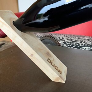 Peut inclure: Un support en bois pour bouteille de vin avec le mot "DUCK" gravé sur le côté. Le support est en bois brun clair et est incliné pour maintenir une bouteille de vin.