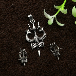 May include: A silver-coloured pendant and two matching earrings featuring a trident design. The pendant has a bail for a chain, and the earrings are smaller versions of the pendant. The trident has three curved prongs and a decorative handle.