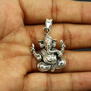 May include: Silver pendant featuring a detailed depiction of the Hindu deity Ganesha. The pendant shows Ganesha sitting with his trunk raised and his hands in a gesture of blessing. The pendant is intricately carved with details on Ganesha's clothing and jewelry.