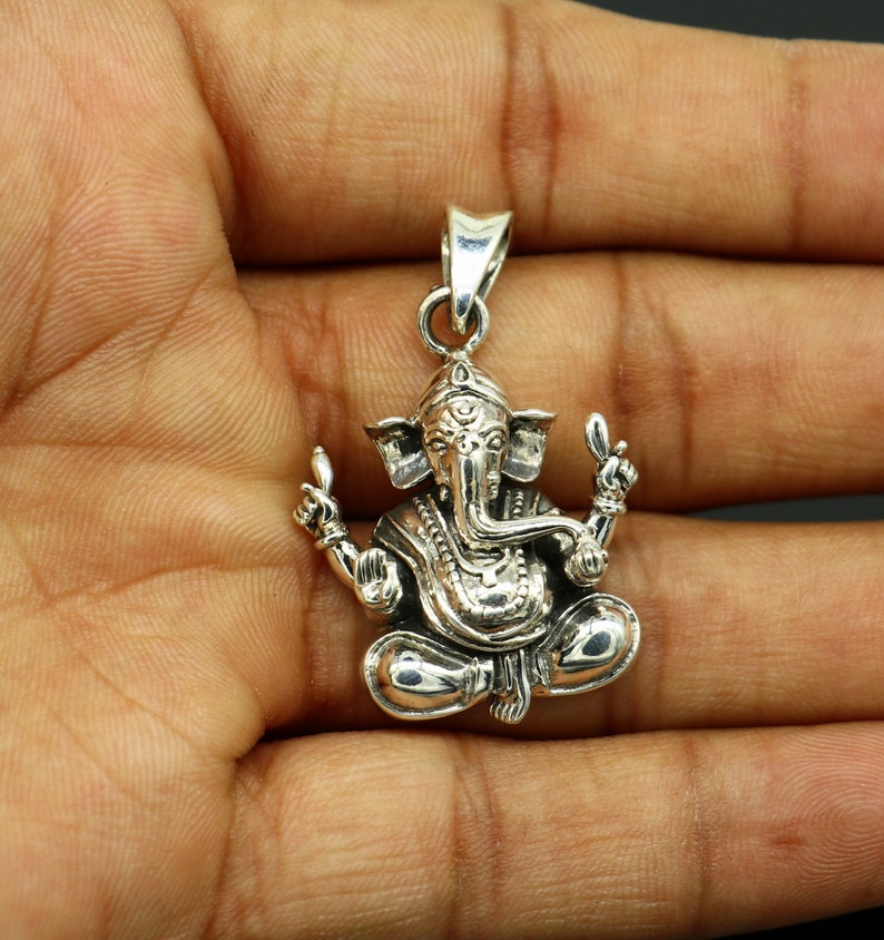 May include: Silver pendant featuring a detailed depiction of the Hindu deity Ganesha. The pendant shows Ganesha sitting with his trunk raised and his hands in a gesture of blessing. The pendant is intricately carved with details on Ganesha's clothing and jewelry.