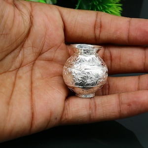 May include: A small, silver, decorative vase with an intricate floral pattern. The vase is about the size of a person's thumb.