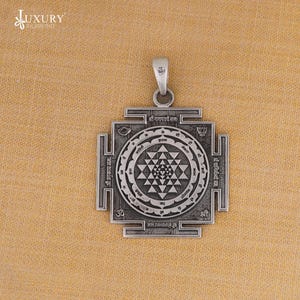 May include: A silver pendant featuring a geometric design and Sanskrit inscriptions. The pendant is square with a central circular pattern and a loop for a chain. Likely crafted from sterling silver, it has intricate details.