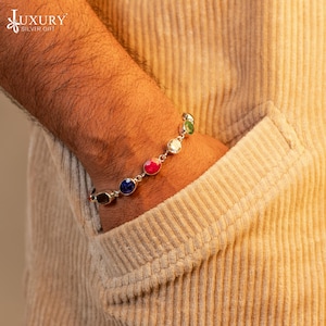 Op de afbeelding: Een zilveren armband met zeven ovale edelstenen in verschillende kleuren, waaronder rood, blauw, groen en wit. De armband zit om een pols.