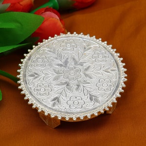 Puede incluir: Un soporte decorativo de plata con una parte superior redonda y tres patas. La parte superior está grabada con un diseño floral.