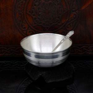 May include: A silver bowl with a spoon, both with intricate designs. The bowl has a smooth, shiny surface and a slightly flared rim. The spoon has a rounded handle with a decorative end.