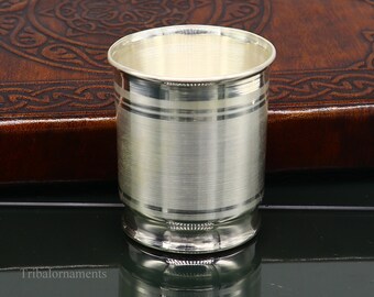 Puede incluir: Una taza de beber de plata con un acabado liso y pulido. La taza tiene un borde ligeramente acampanado y una base redondeada. Está sentada sobre una superficie oscura.